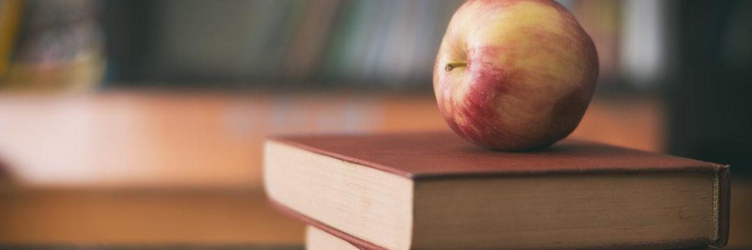 Apple sitting on a pile of books on a classroom desk.