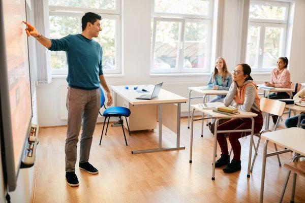 A teacher points to the board at the front of a classroom as students at desks listen.